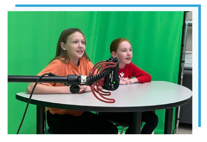 Two students sitting with a mic and a green screen preparing to record a video